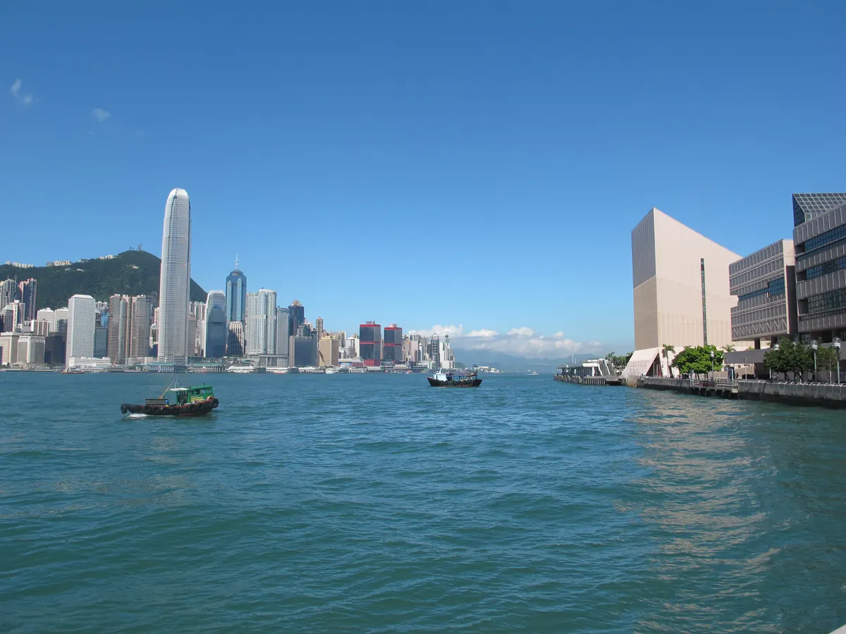 Victoria Harbour in clear morning light from the Tsim Sha Tsui waterfront: a boat on the blue water and the Hong Kong Island skyline, with the tall Two IFC tower, across the harbour.