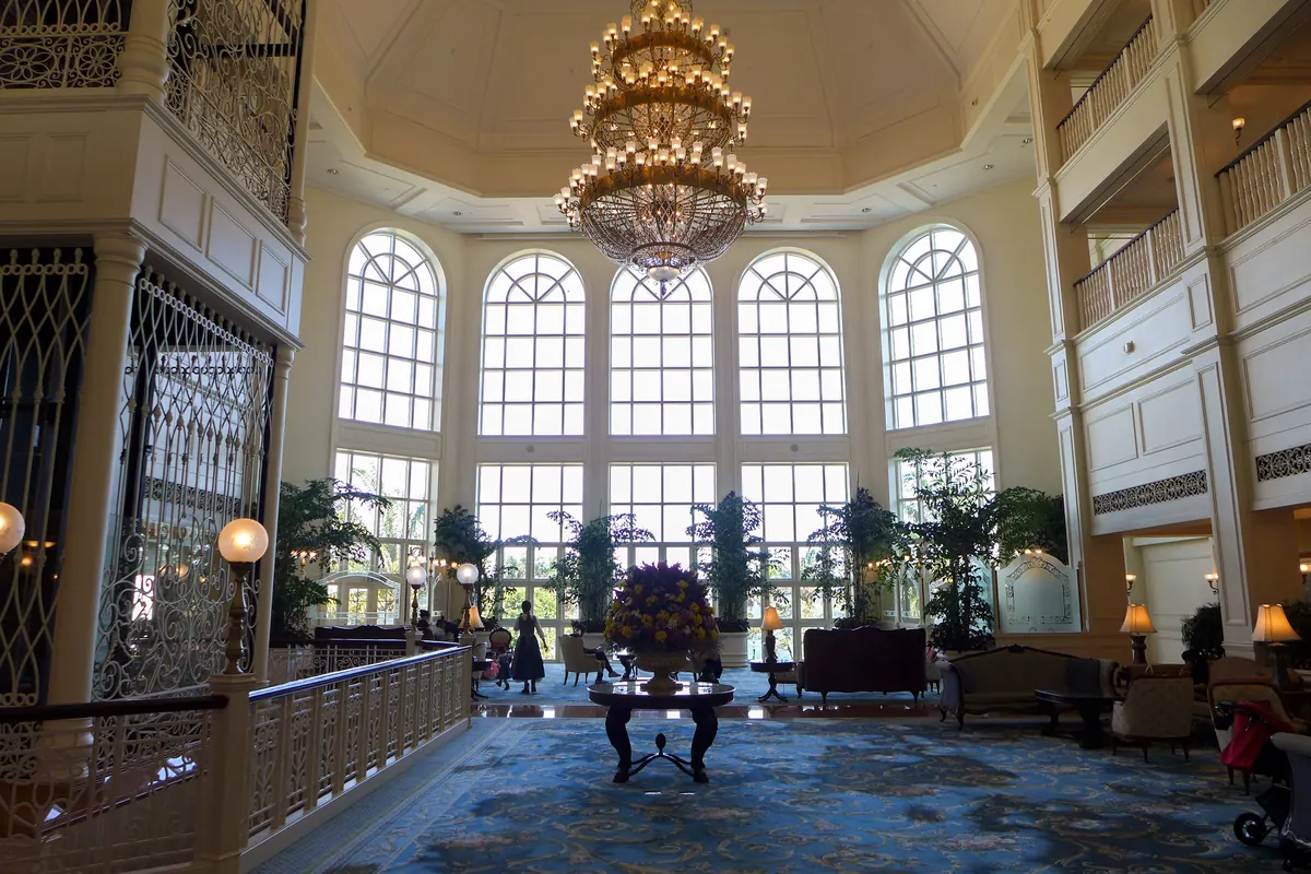 The grand lobby of the Hong Kong Disneyland Hotel: a huge crystal chandelier under a high ceiling, tall arched windows full of daylight and a blue patterned carpet.