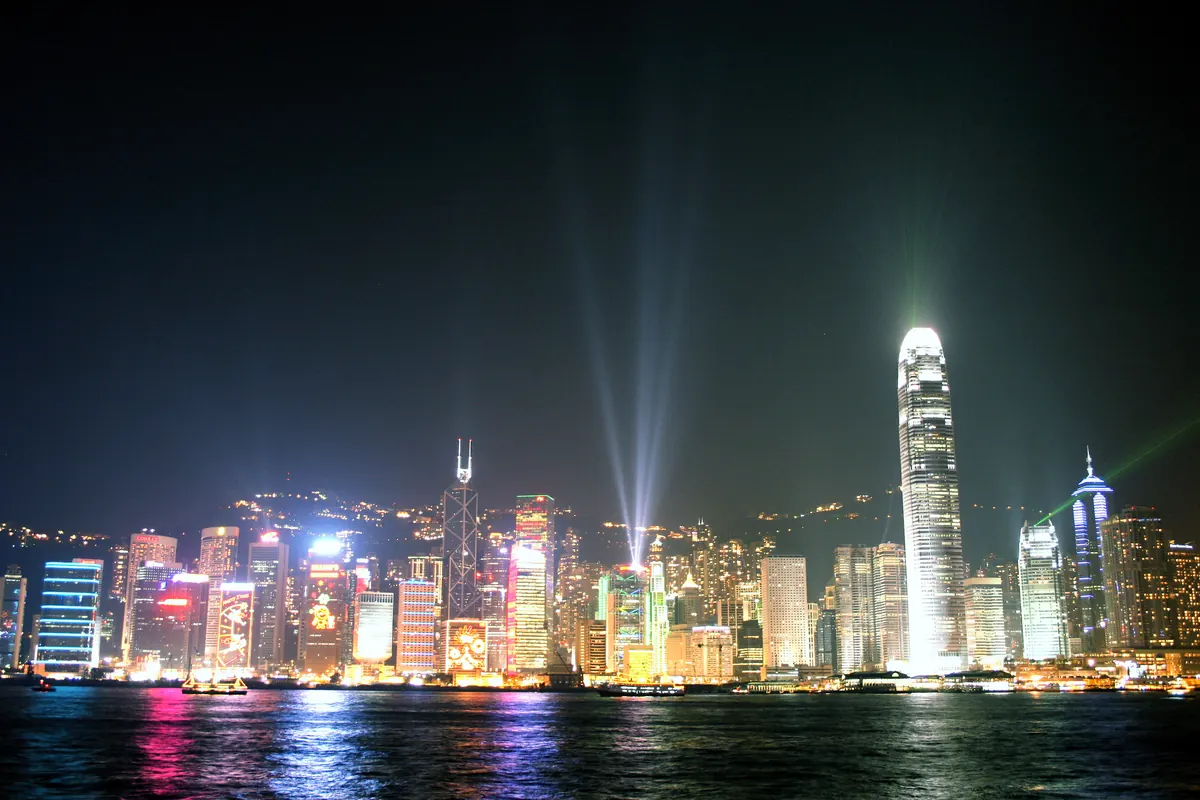 The Hong Kong Island skyline lit in colour across Victoria Harbour at night, with beams of light from the Symphony of Lights show rising above the towers, seen from the Tsim Sha Tsui waterfront.