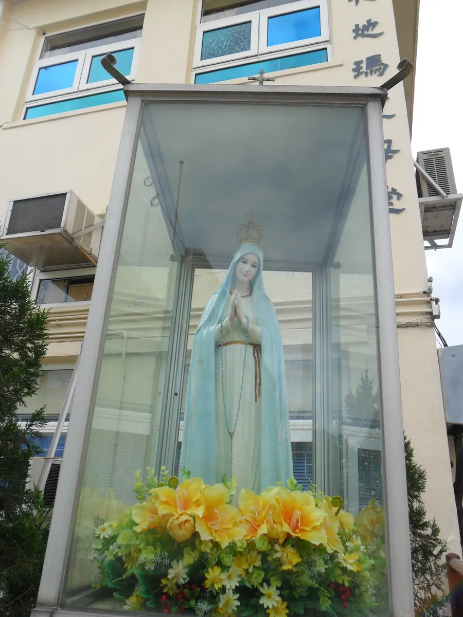 The statue of Our Lady of Fátima, in a blue veil and crown with her hands joined in prayer, standing in a glass shrine ringed with yellow flowers outside Our Lady of Fátima Church on Cheung Chau.